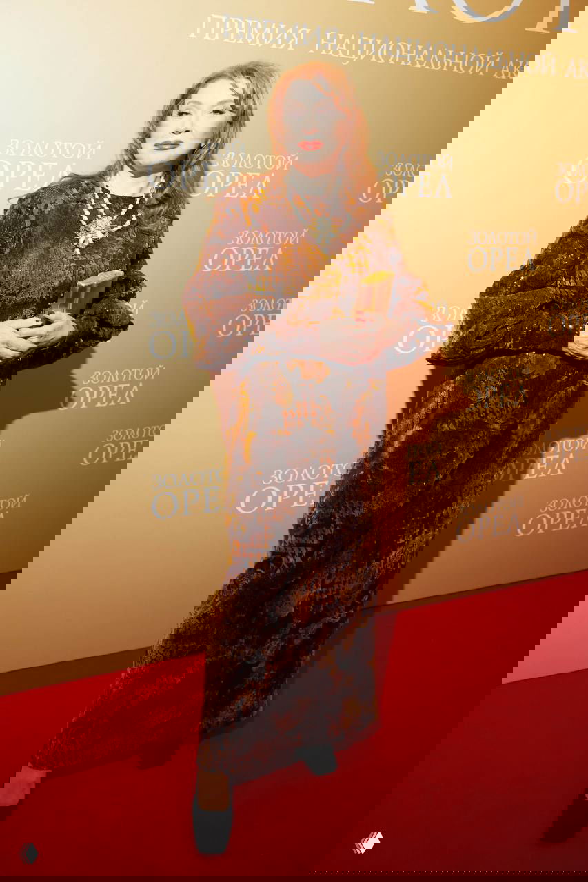 Вера Сотникова в длинном темно-коричневом бархатном платье с украшением на шее держит статуэтку и позирует на красной дорожке «Золотой орёл».