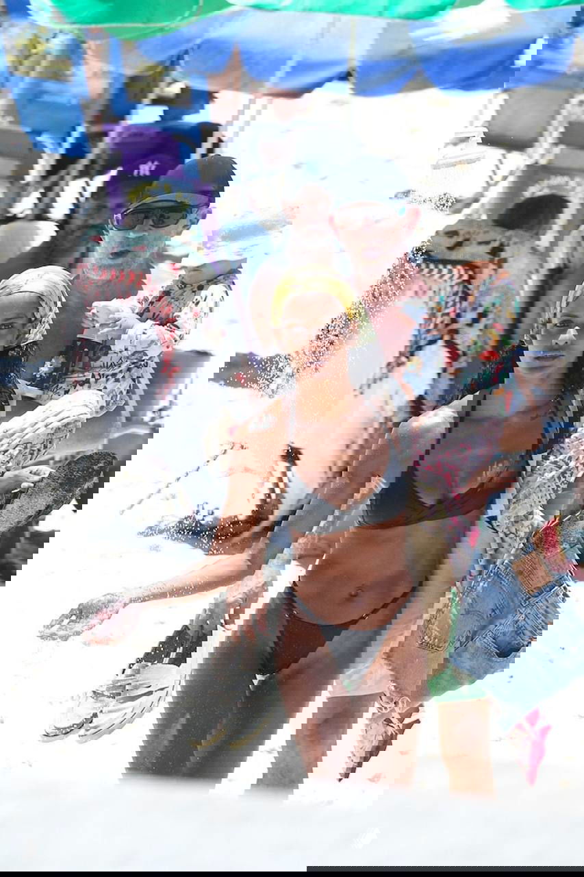 Женщина в чёрном купальнике идёт по пляжу вперёд, мужчина держит ребёнка и несёт обувь; вокруг пляжная экипировка и зонты.