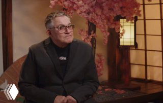 Мужчина средних лет в тёмном пиджаке сидит в интерьерной студии на фоне декоративных розовых цветов и тёплого мягкого освещения, кадр интервью