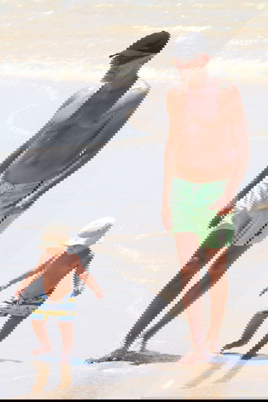 Мужчина в тёмной кепке идёт по кромке воды рядом с маленьким ребёнком в ярких шортах; следы ног на влажном песке.