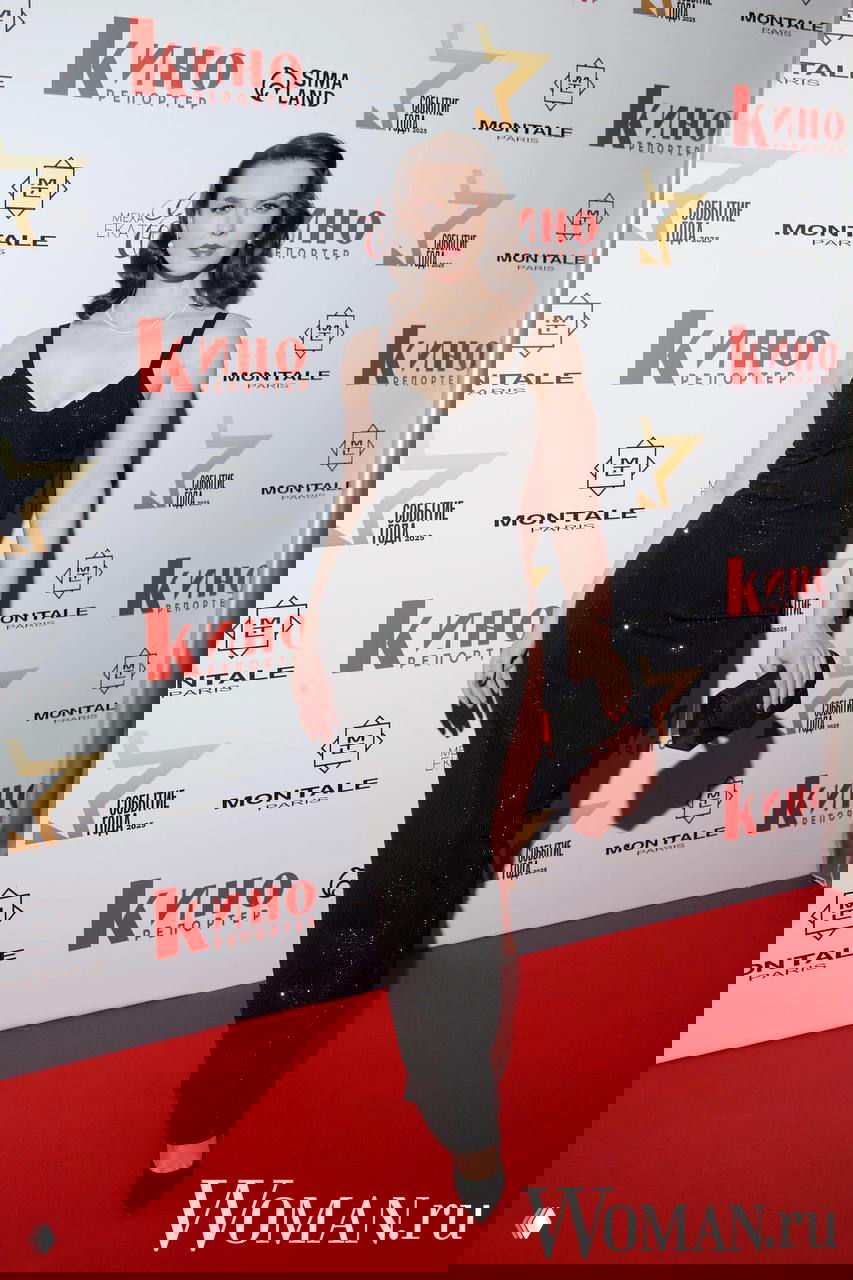 Женщина в чёрном длинном платье с открытыми плечами и жемчужным украшением, полный рост на красной дорожке у баннера мероприятия.