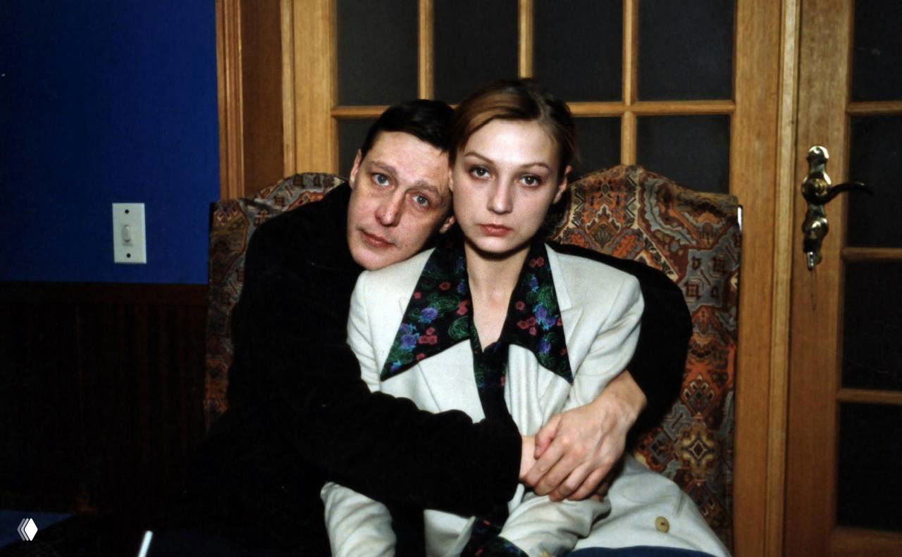 Фотография в интерьере: мужчина обнимает женщину на кресле у деревянной двери; видно серьёзные лица и домашняя обстановка.