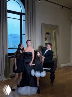 Групповой семейный кадр в светлом интерьере: мужчина у рояля и несколько девушек в вечерних нарядах на фоне большого окна и города.