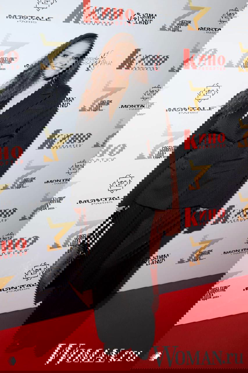 Женщина в двубортном тёмном костюме и широких брюках позирует на красной дорожке перед фотостеной «Кино Репортер».