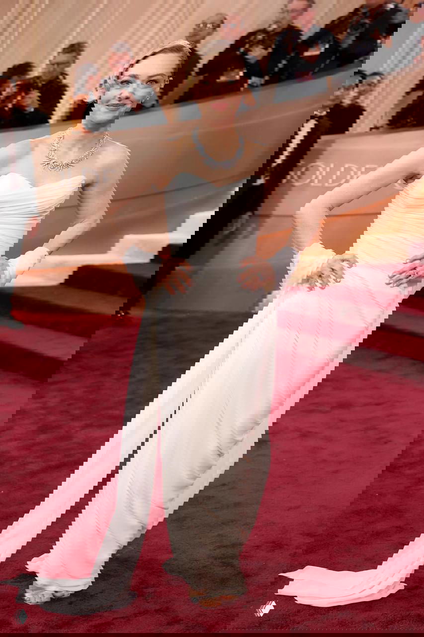 Женщина в белом классическом платье со струящимся шлейфом на красной дорожке, вокруг фотографы и на фоне виден фотоколл события