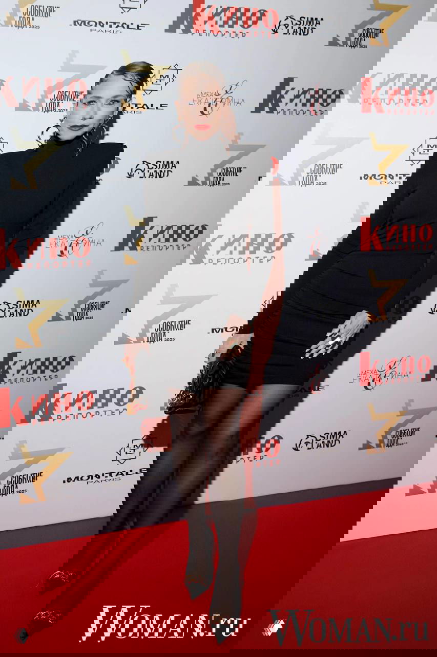 Женщина в мини-платье чёрного цвета с длинными рукавами и аксессуарами, полный рост на красной дорожке у бренд-стены мероприятия.