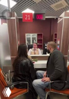 Пара в кабинете ЗАГСа: женщина и мужчина сидят напротив регистратором за столом; на заднем плане видно табло, кресла и потолок.