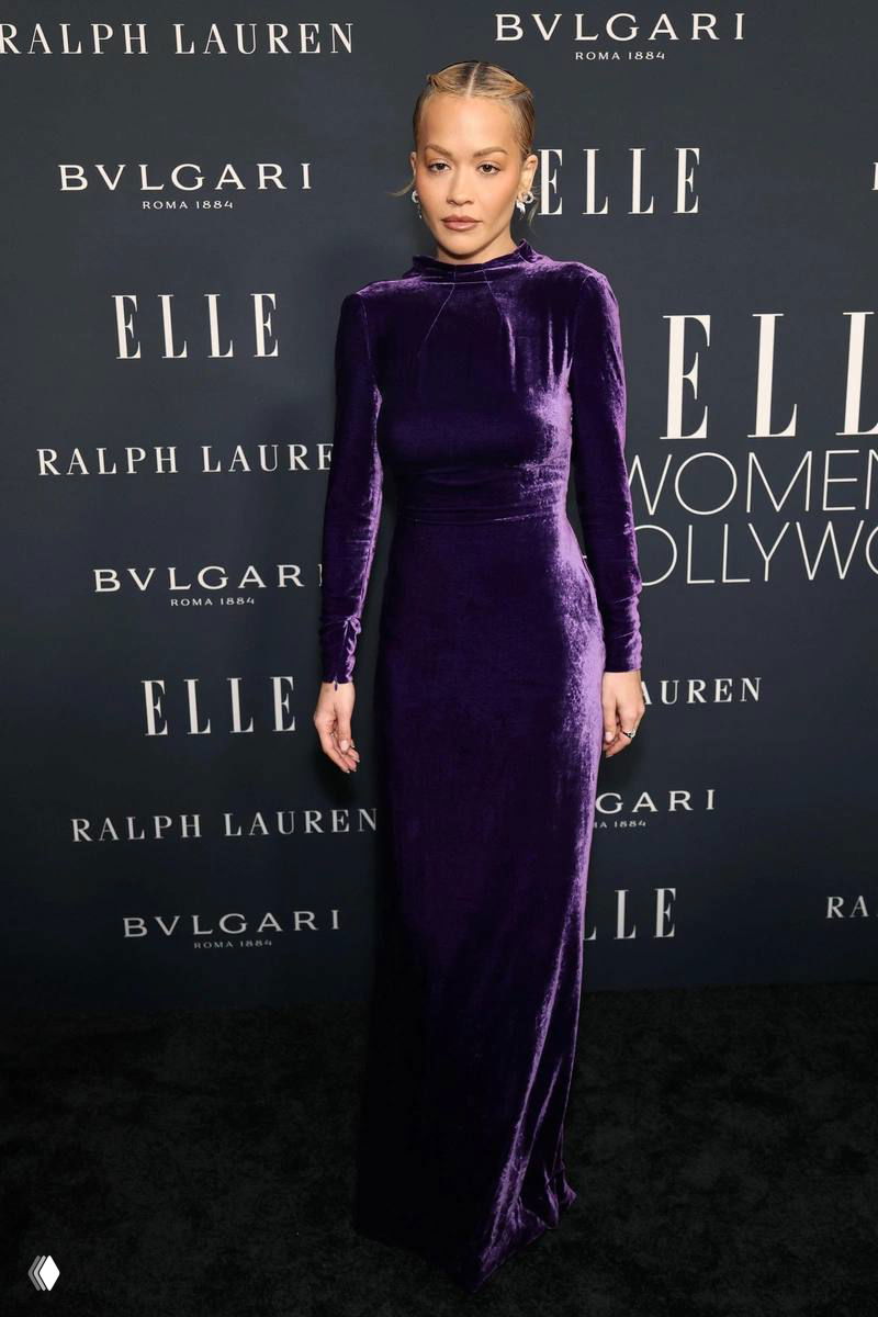 Рита Ора на церемонии ELLE Women in Hollywood в длинном бархатном фиолетовом платье, снимок в полный рост на фоне баннера ELLE и спонсоров.