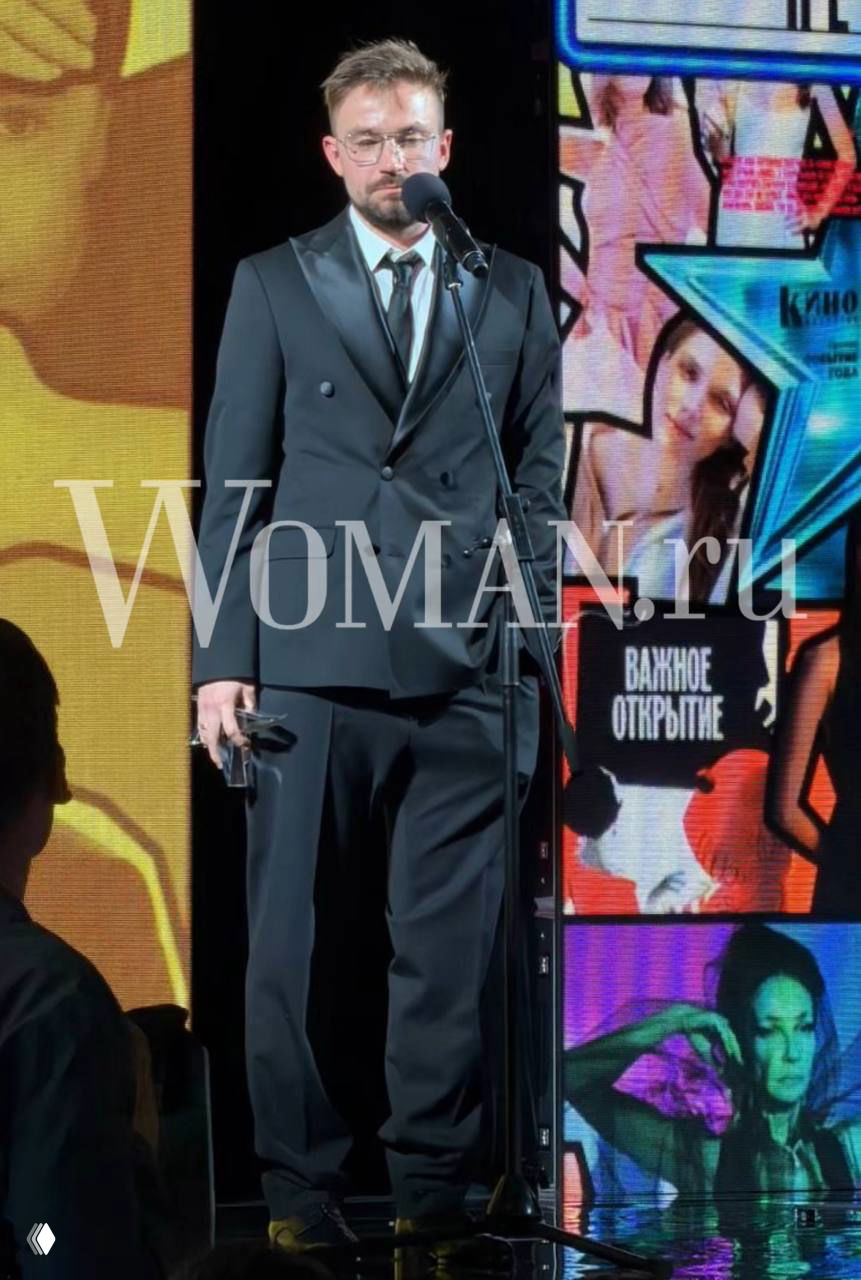 Александр Петров в костюме стоит у микрофона на сцене церемонии «Кино-репортер»; на заднем плане яркий экран и логотип Woman.ru.