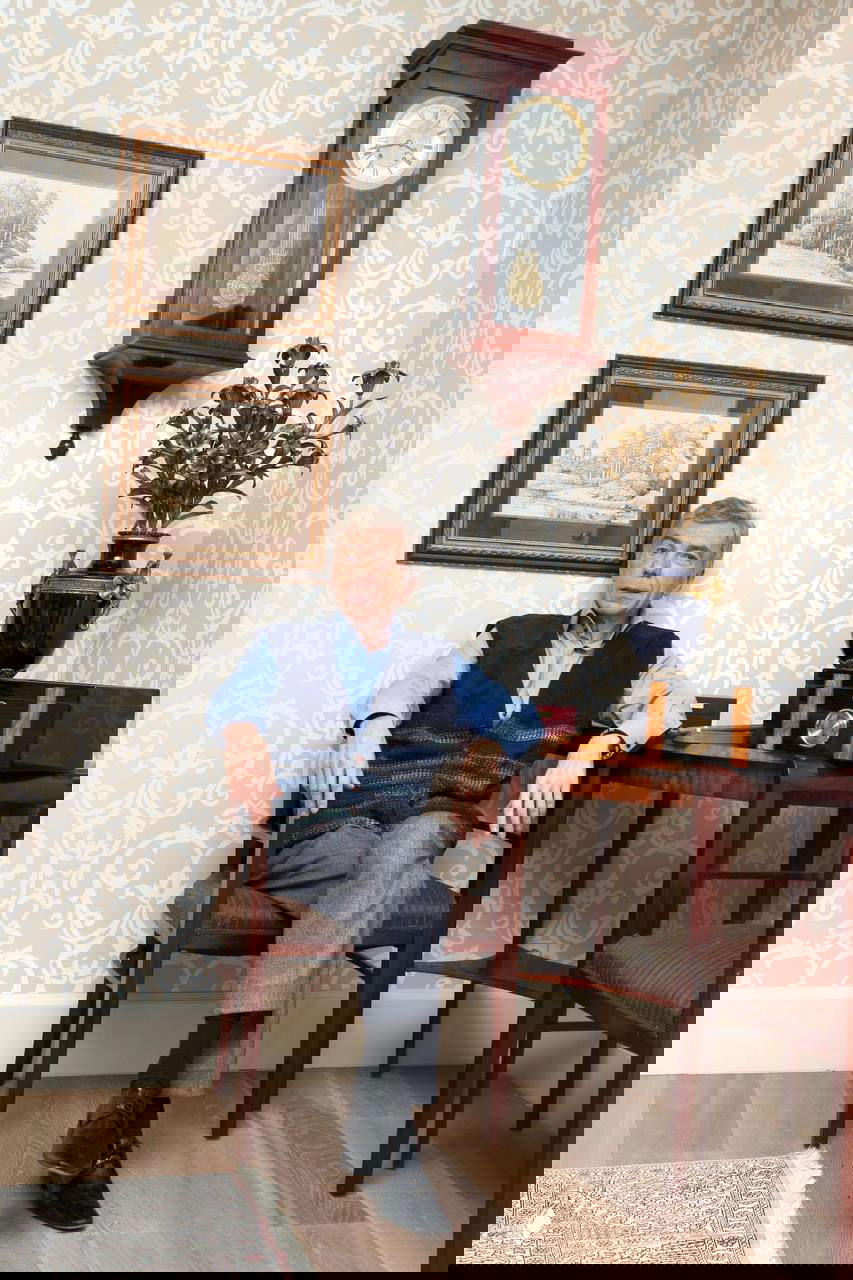 Юрий Николаев в интерьерной фотосессии: телеведущий сидит в кресле в гостиной рядом с настенными часами и декоративной вазой, домашняя обстановка.