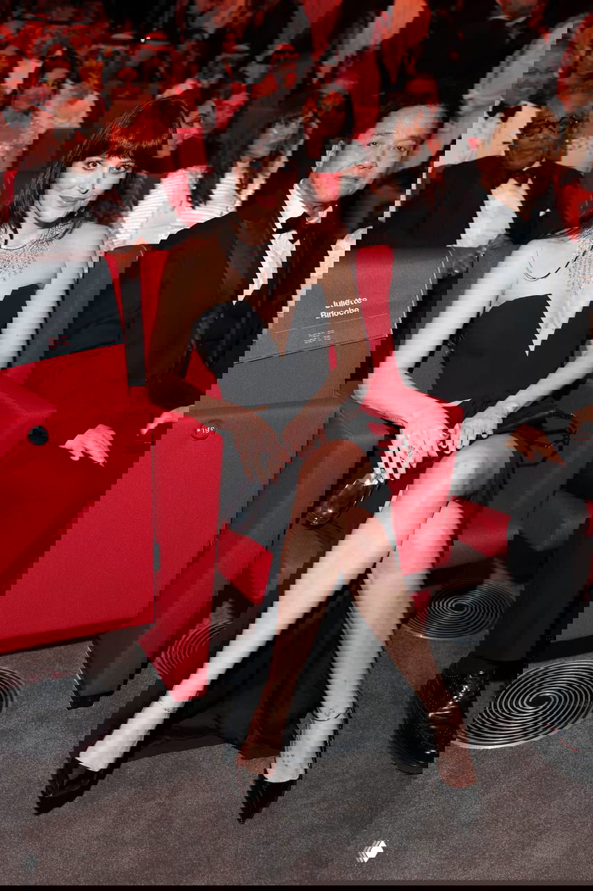 Актриса на красной дорожке в сидячем кадре: черное платье с глубоким декольте, крупное ожерелье и зрители в зале рядом.