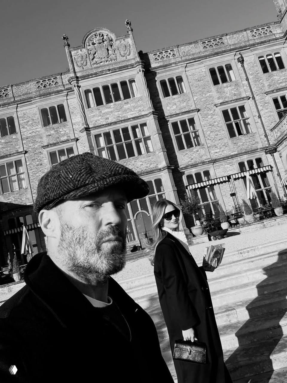 Чёрно‑белый широкоугольный селфи‑кадр: мужчина и женщина на ступенях перед фасадом старинного дома, композиция подчёркивает масштаб архитектуры и семейный момент