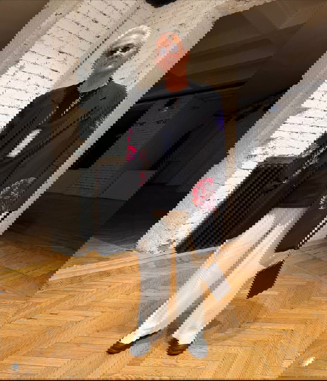 Олег Меньшиков в чёрном пиджаке с цветочными аппликациями, тёмной футболке и светлых свободных брюках — элегантный вечерний образ на фоне галерейного интерьера.