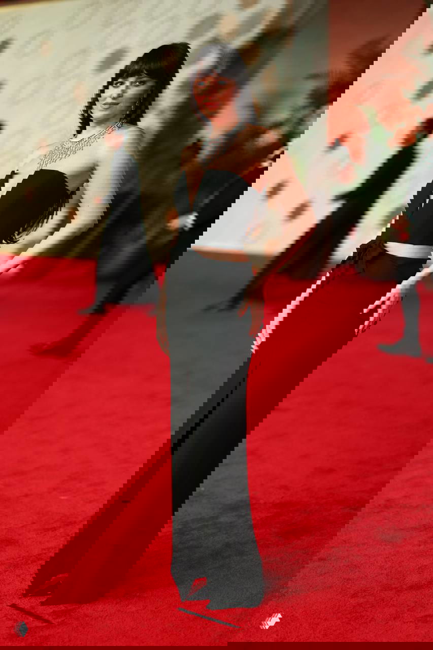 Полноразмерный снимок на красной дорожке: черное приталенное платье с вырезами на талии, актриса позирует на фоне гостей фестиваля.