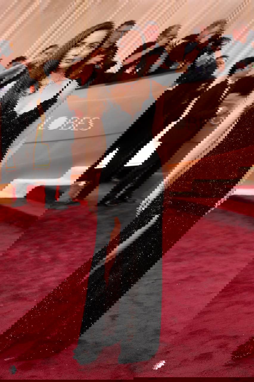 Женщина в чёрном платье с кружевной отделкой и высоким разрезом, полный рост на красной дорожке, вокруг — фотографы и гости мероприятия.