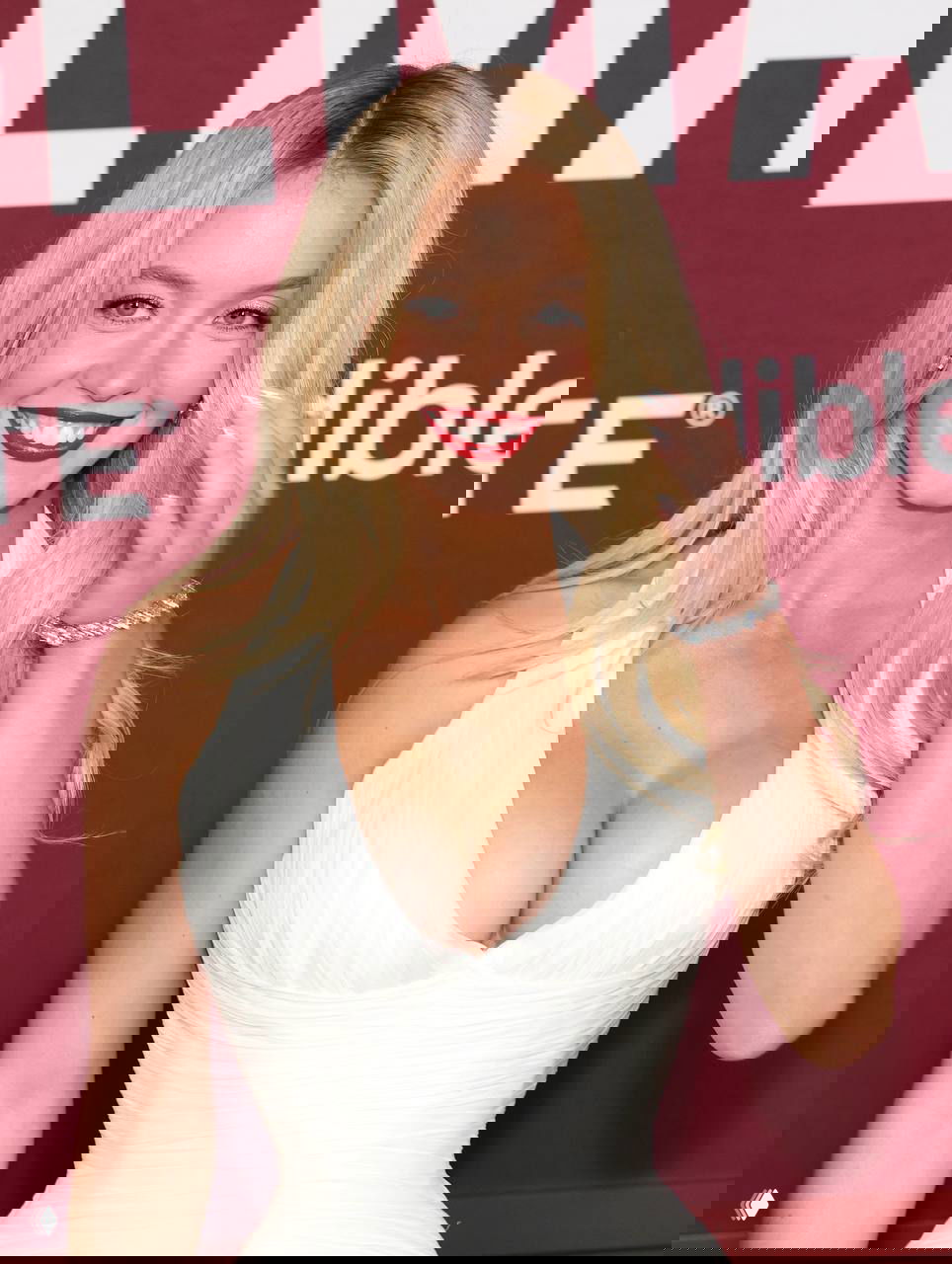 Крупный план женщины в белом платье на фоне постера премьеры: красная помада, украшения и прическа на фотоколле в Лос‑Анджелесе.