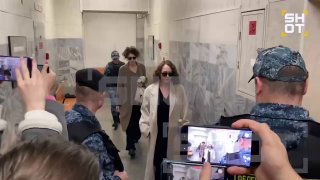 Женщина в светлом пальто идёт по коридору здания в сопровождении сотрудников правопорядка и журналистов с камерами, обстановка похожа на судебное заседание