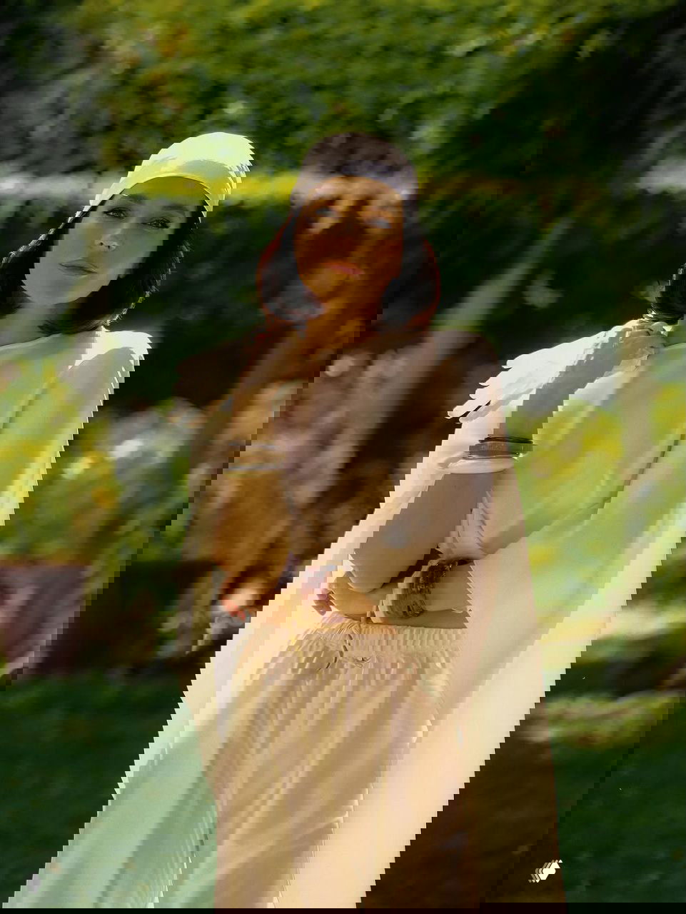 Фотография в полный рост: женщина в светлом костюме на фоне зелёного сада, солнечные блики подчёркивают текстуру ткани и натуральный макияж.