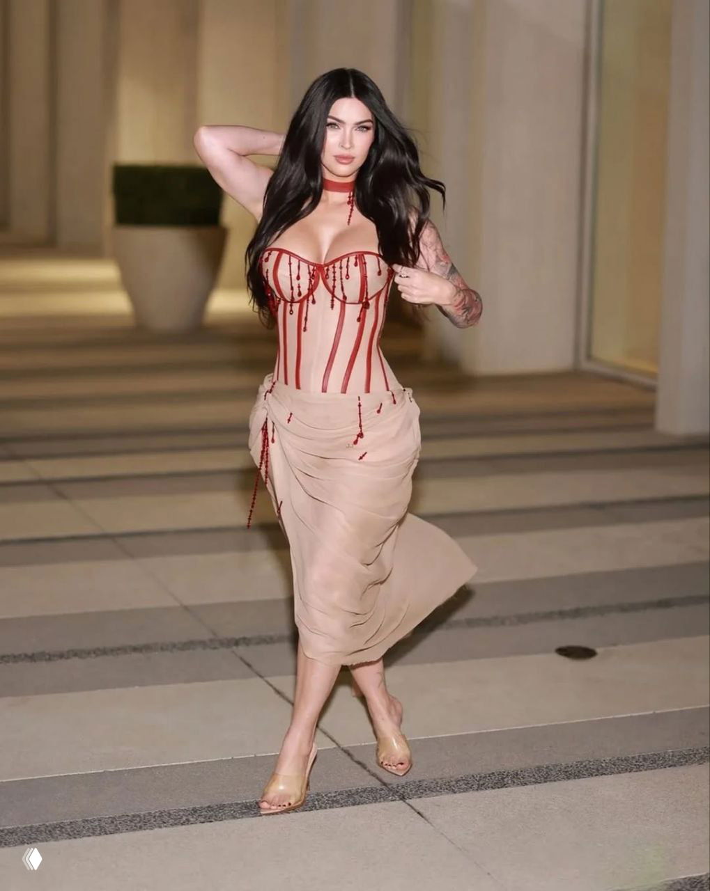 Женщина в бежевом корсете с красными вертикальными элементами и прозрачной юбкой, идёт по дорожке на вечернем показе.