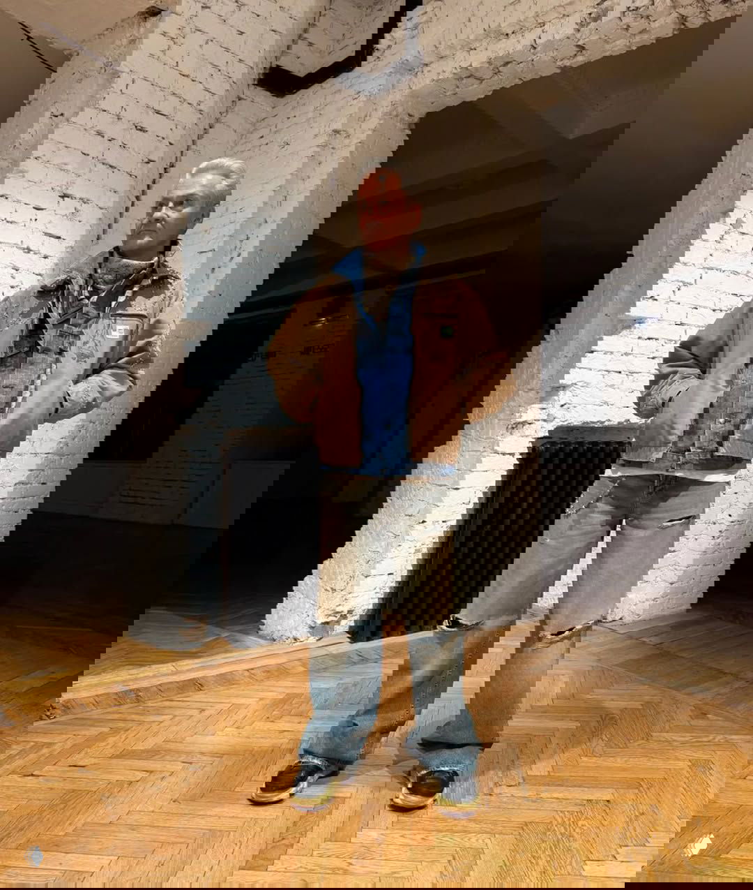 Слоистый деним‑лук: джинсы с потертостями, джинсовая рубашка и коричневая куртка поверх — мужской расслабленный стиль на паркетном полу.