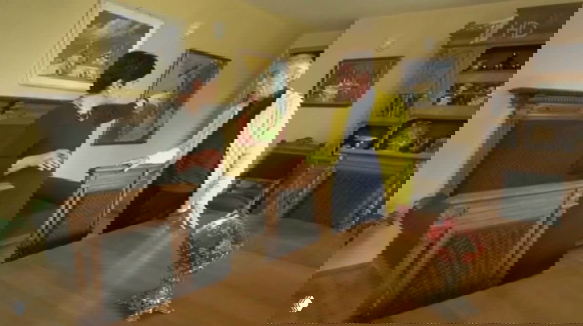 Николаев в беседе с женщиной в столовой с деревянной мебелью и картинами на стенах: интерьер с предметами антиквариата и декором.