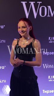 Молодая гостья в чёрном платье и украшениях улыбается на фоне пресс-волла с логотипом Woman.ru во время праздничного события издания.
