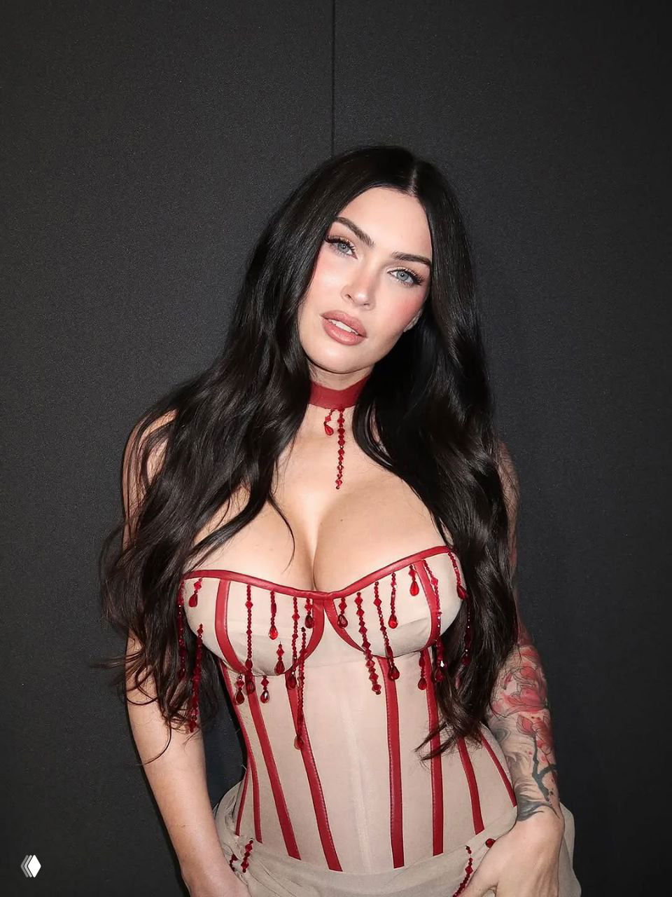 Женщина в бежевом корсете с красными бусинами и имитацией капель крови, длинные чёрные волосы, портрет на тёмном фоне.