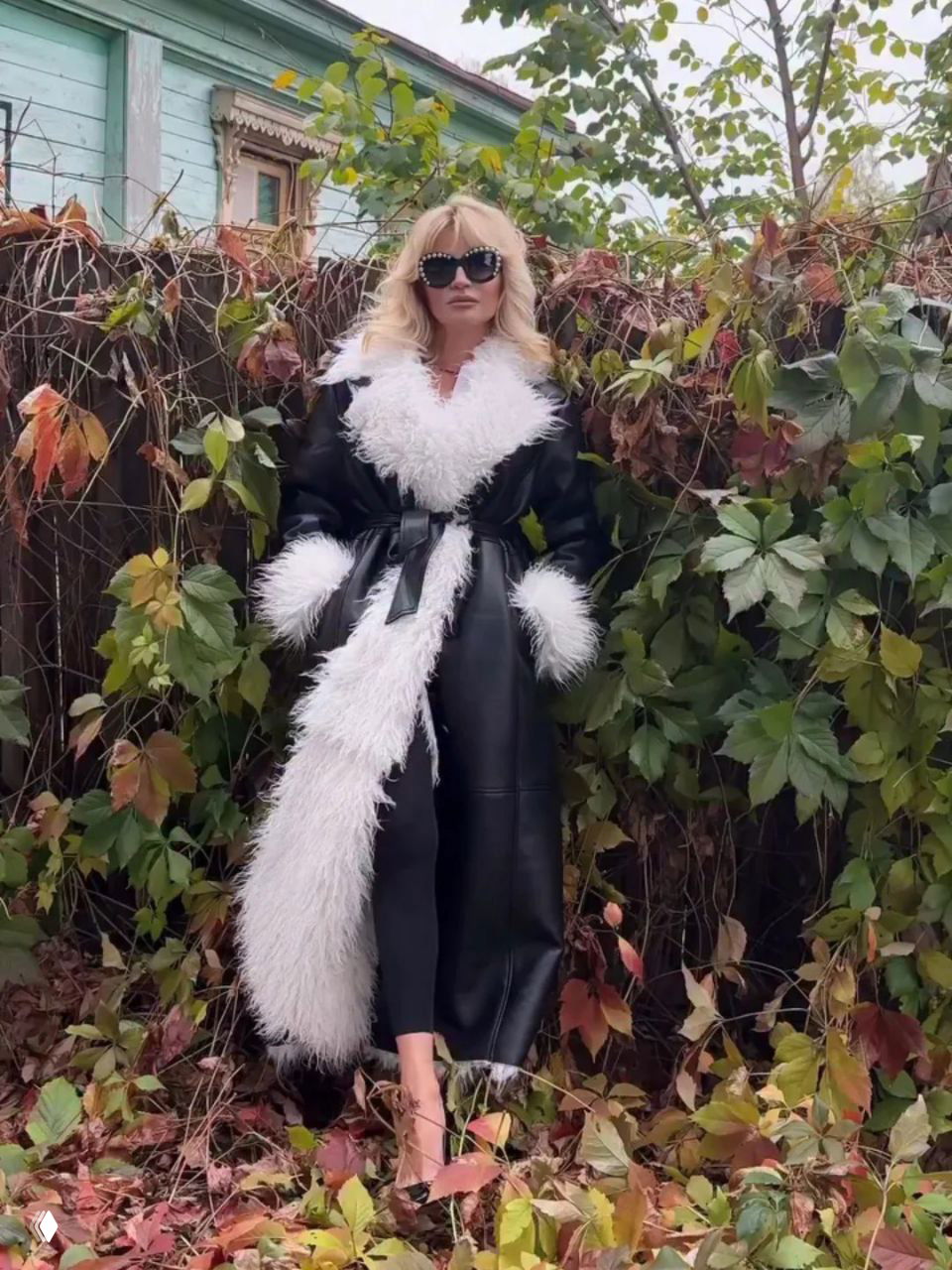 Женщина в длинном чёрном кожаном пальто с белой пушистой отделкой позирует на фоне осенней лианы и опавшей листвы, стильный городской кадр.