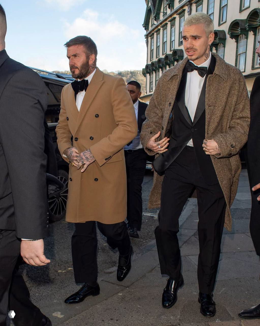 Дэвид Бекхэм в светло-коричневом пальто и сын Круз в смокинге идут по тротуару в сопровождении охраны на свадебном мероприятии