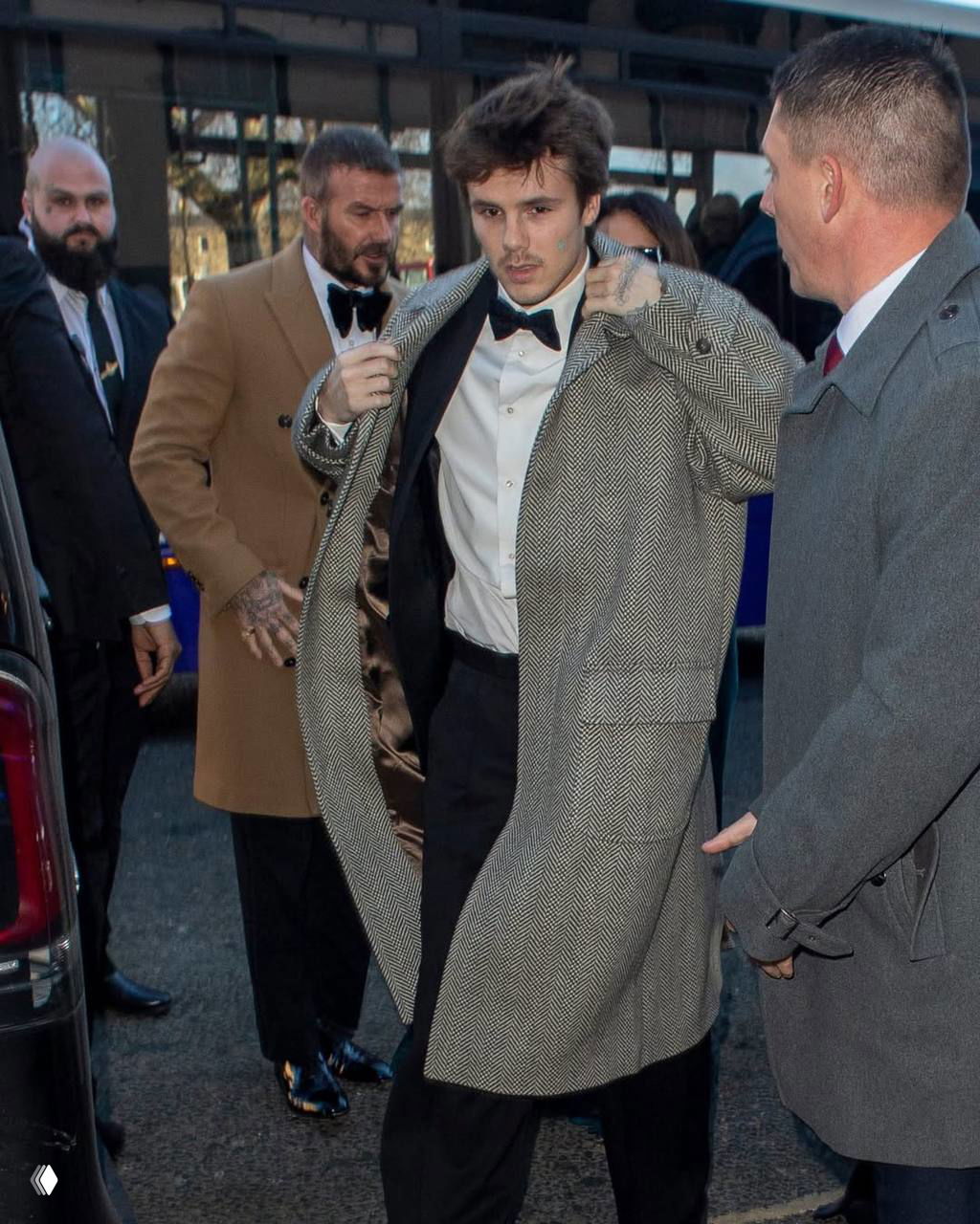 Ромео Бекхэм в смокинге и длинном пальто поправляет одежду при выходе из автомобиля на фоне сопровождающих — момент прибытия на свадьбу