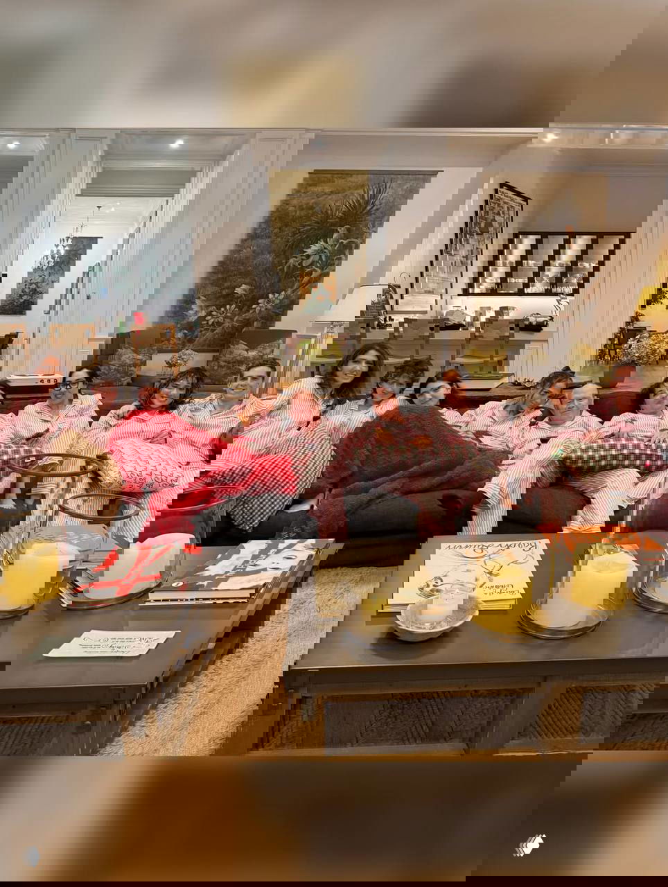 Несколько человек в одинаковых полосатых пижамах сидят на длинном диване в гостиной; на переднем плане журнальные столики со свечами и книгамии.