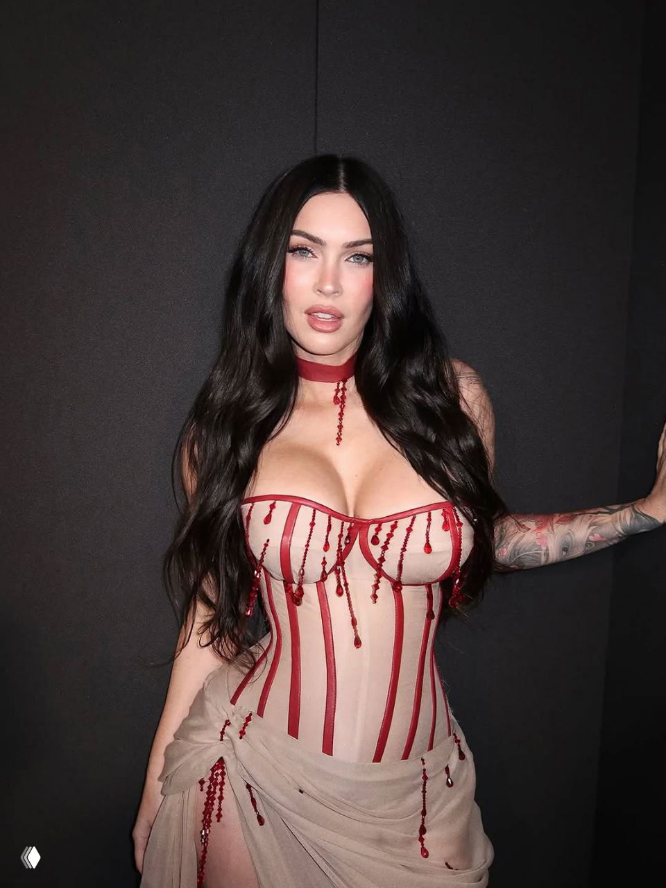 Крупный план женщины в бежевом корсете с красными деталями, выраженный макияж и длинные чёрные волосы на тёмном фоне.