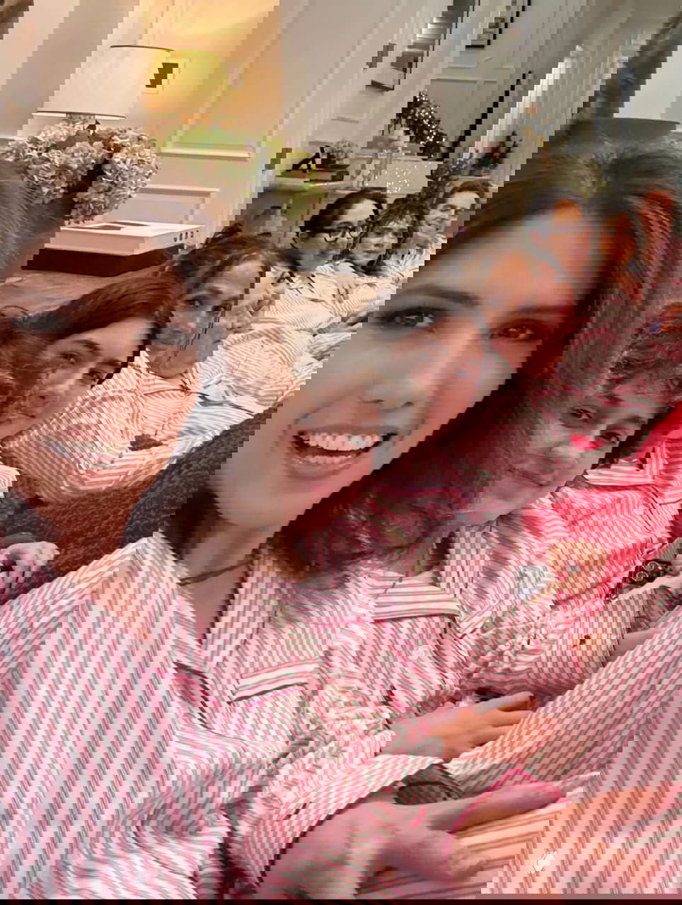 Групповое селфи: несколько людей в полосатых пижамах сидят вплотную на диване на фоне рождественского декора и гирлянд, улыбки и тёплая атмосфера.