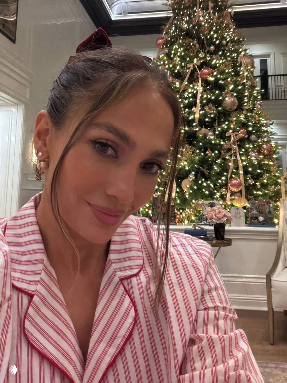 Портретная фотография женщины в полосатой пижаме на фоне высокой украшенной рождественской ёлки с золотыми и розовыми игрушками и гирляндами.