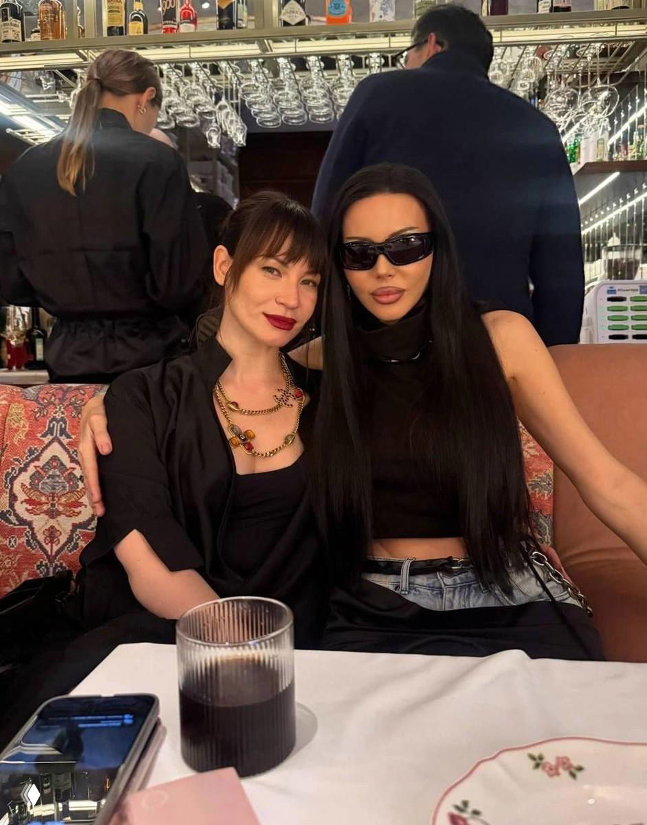 Две посетительницы в чёрной одежде сидят за столом ресторана; на переднем плане сервировка и стакан с напитком