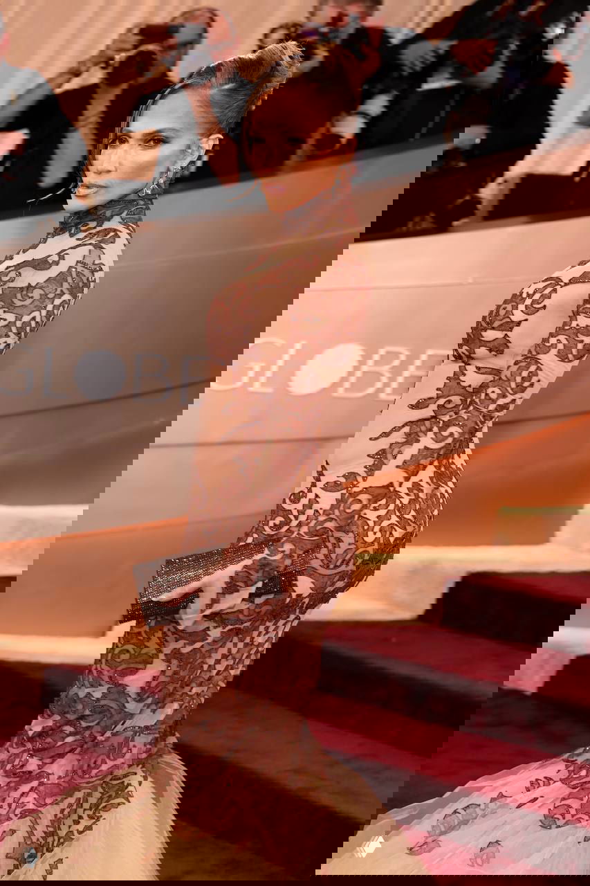 Боковой портрет Дженнифер Лопес в прозрачном платье с коричневой вышивкой на церемонии «Золотой глобус», клатч в руке, фотографы позади.