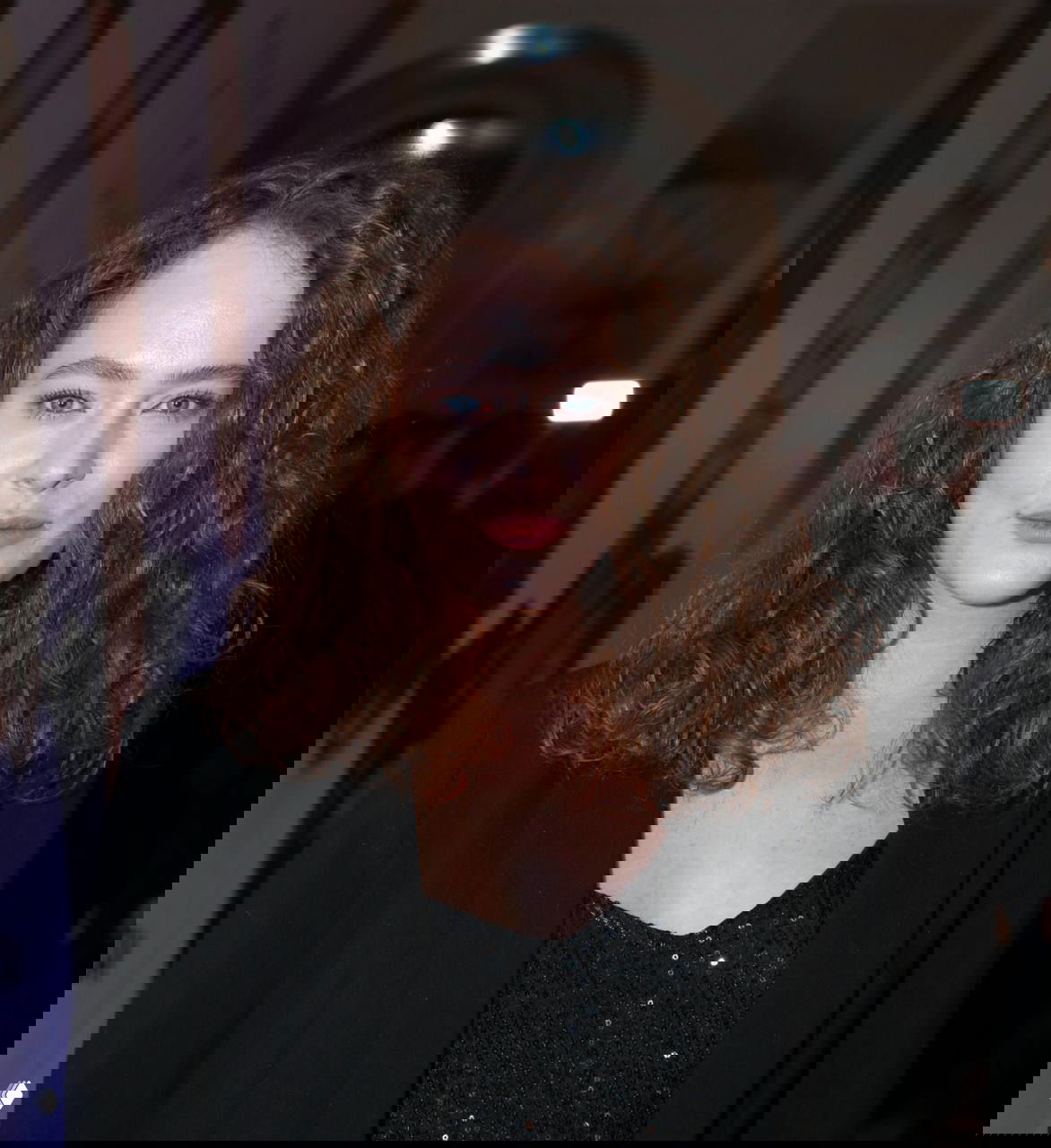 Крупный план женщины в вечернем образе с кудрявыми волосами и сдержанным макияжем на фоне светского мероприятия.