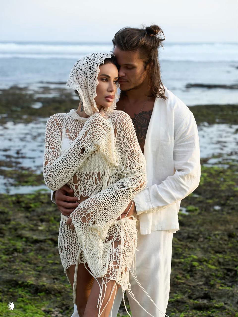 Айза и её новый партнёр Степан Кузьменко обнимаются на каменистом побережье в светлой одежде; виден морской прибой и романтическая атмосфера.