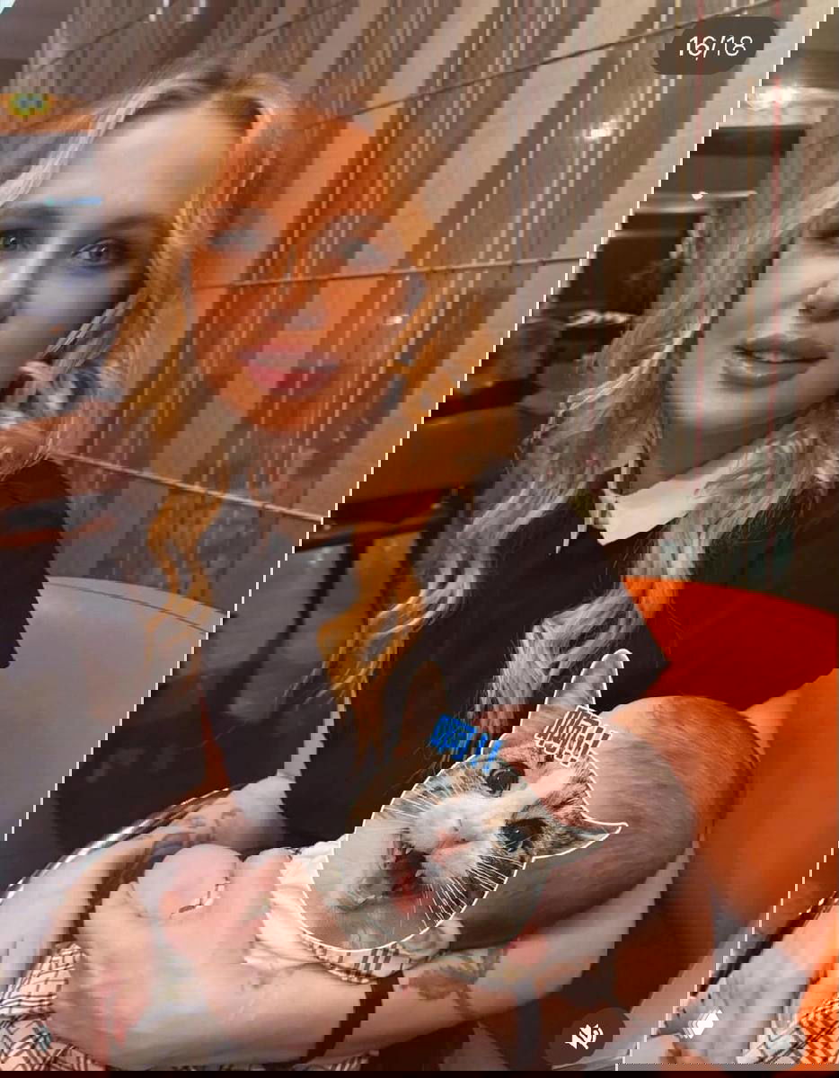 Женщина держит новорождённого ребёнка на руках в кресле; лицо младенца закрыто наклейкой с изображением кота, семейный кадр в интерьере.