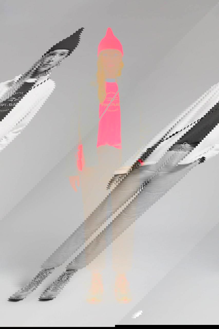 Молодая женщина в светлой флисовой куртке, красной шапке и бежевых утеплённых брюках на сером фоне — повседневная тёплая экипировка из натуральных оттенков.