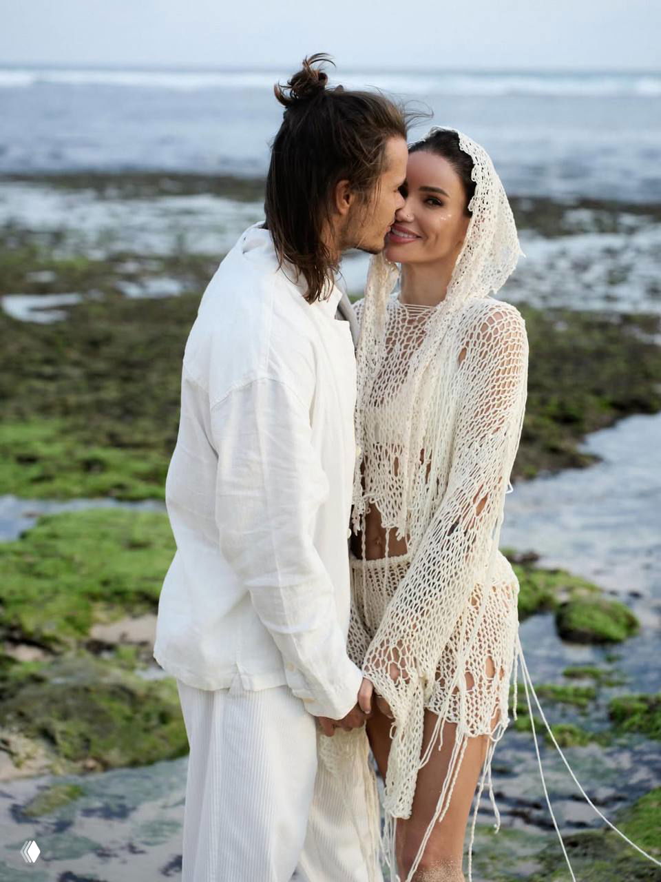 Айза и Степан улыбаются и держатся за руки у воды в белых нарядах; на заднем плане видны отмели и морские скалы, крупный план пары.