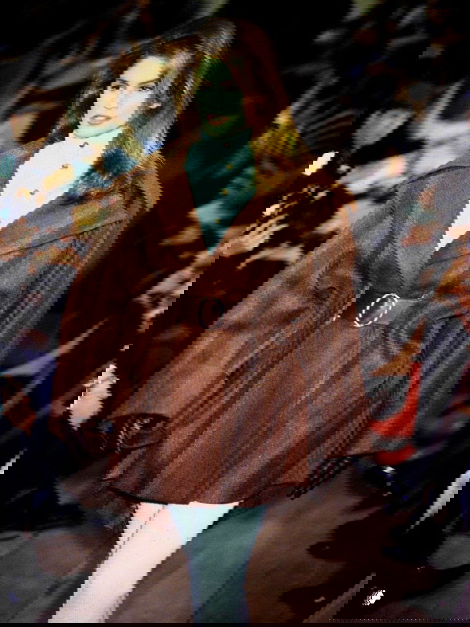 Архивный кадр модели на подиуме в коричневом пальто с широким поясом и белых брюках, показная съёмка с публикой и ламповым светом.