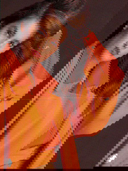 Ретро‑портрет модели в оранжевой рубашке в расслабленной позе, кадр подчёркивает макияж, цветовую палитру и гламурную эстетику 1990‑х.