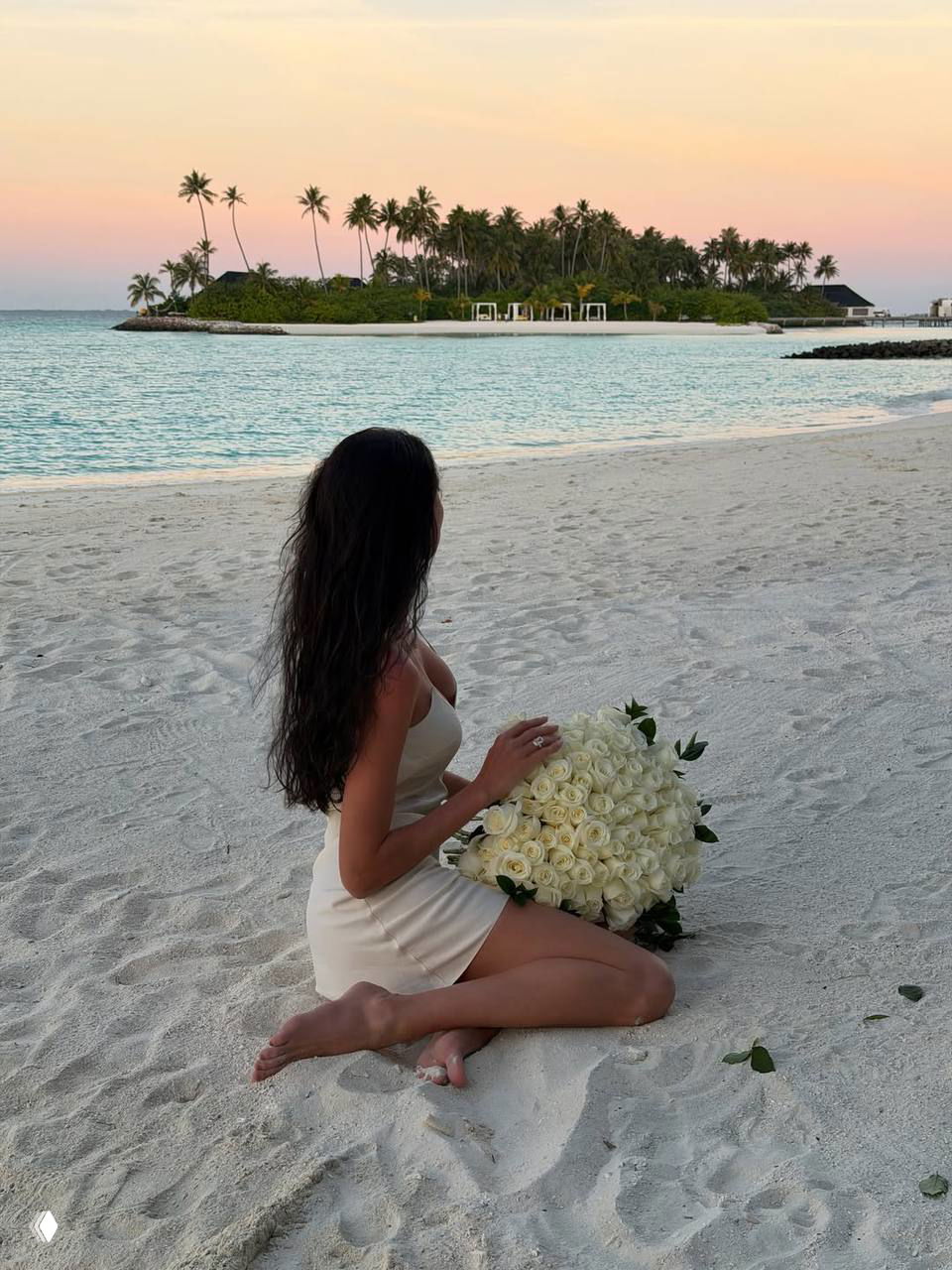 Женщина в белом платье сидит на песчаном берегу и держит большой букет белых роз; на заднем плане — остров с пальмами и море при закате.