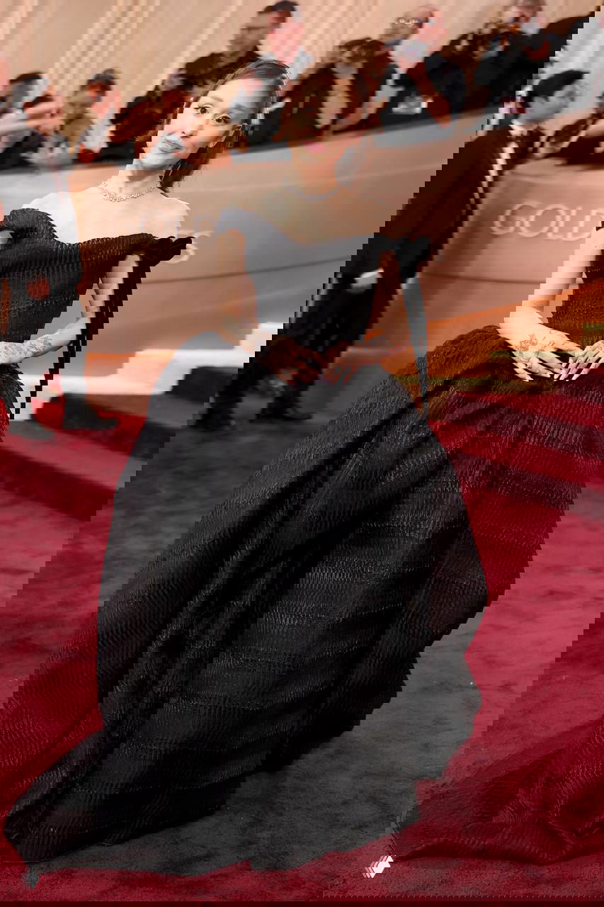 Женщина в пышном чёрном вечернем платье с открытыми плечами стоит на красной дорожке, авторы снимков и гости вокруг.