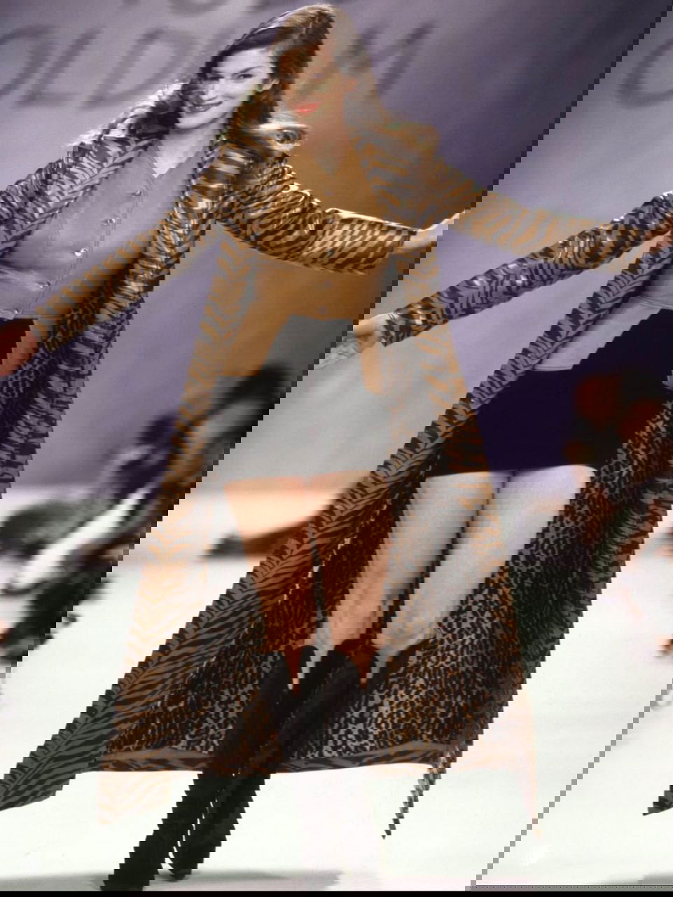 Фотография с показа: модель в длинном тигровом пальто и мини‑юбке, динамичный кадр на подиуме с публикой и ретро‑атмосферой.