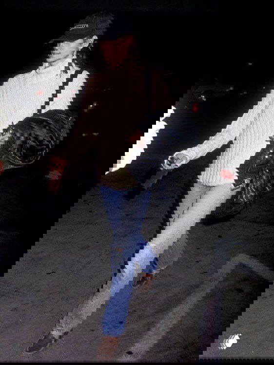 Архивная уличная фотография модели в джинсах, светлой рубашке и кепке, спонтанный снимок в ночном городском пространстве с неформальной позой.