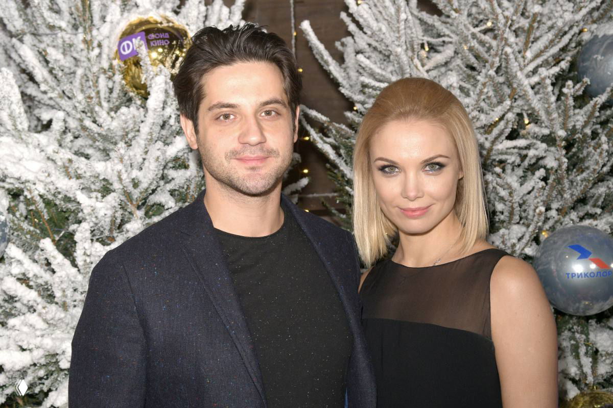 Два человека в вечерней одежде позируют на фоне украшенной заснеженной ёлки с гирляндами и шарами на светском праздничном мероприятии.