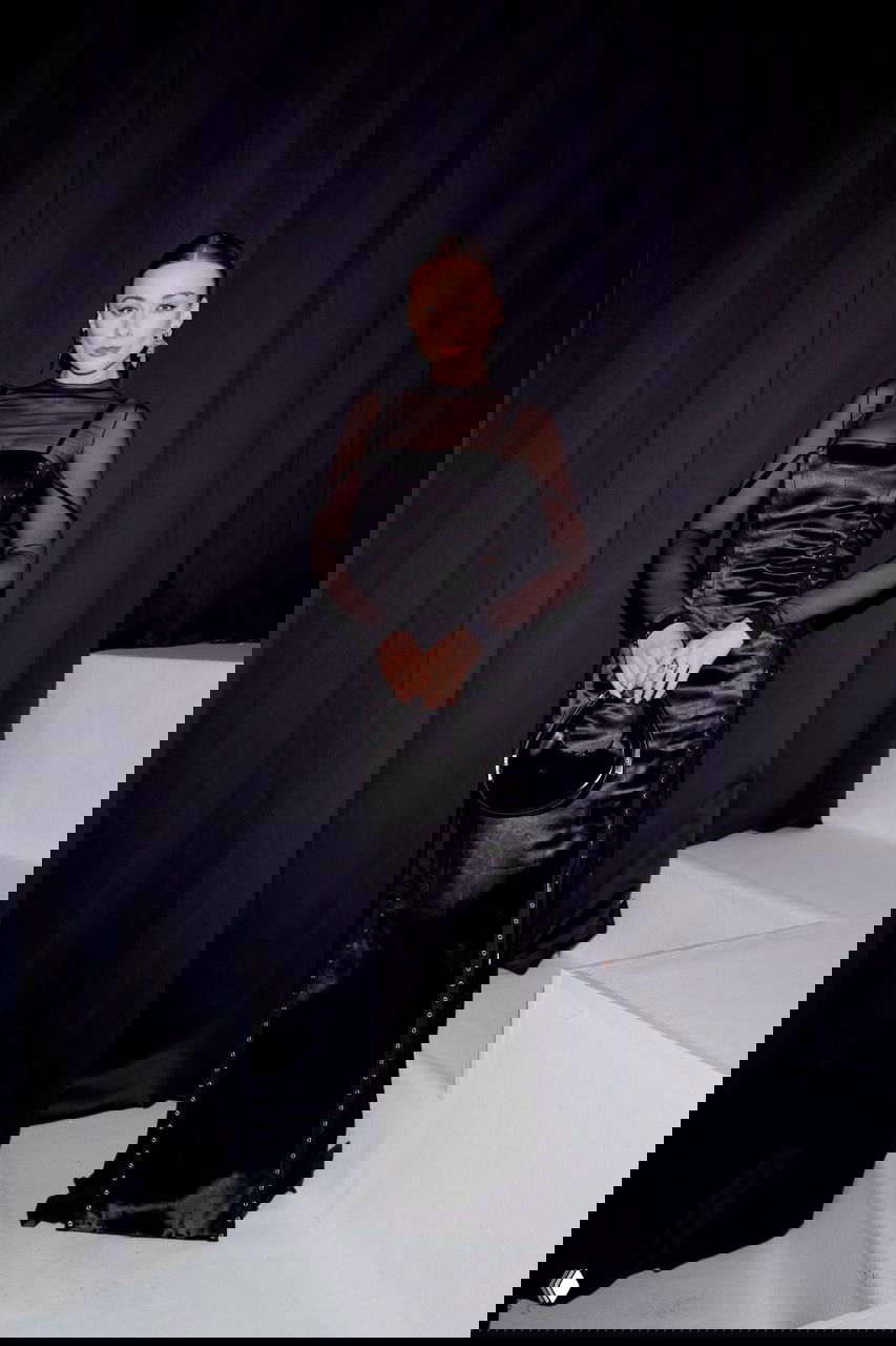 Гостья в чёрном атласном платье с прозрачными рукавами стоит у тёмной шторы — полный рост, стильный вечерний образ для гала-ужина.