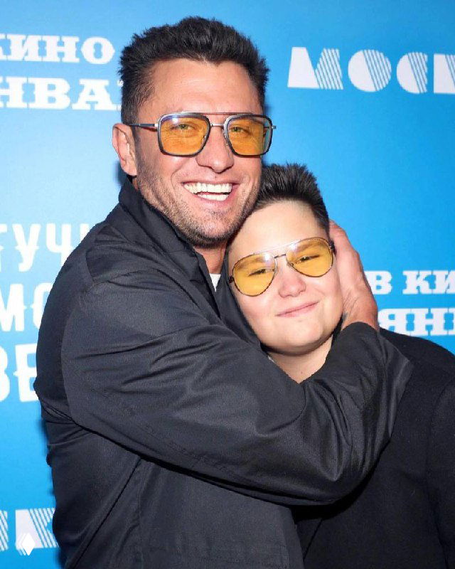 Мужчина и мальчик обнимаются на фоне баннера на светском мероприятии: мужчина улыбается, ребёнок в жёлтых очках.